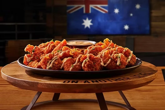 Platter of crispy boneless fried chicken pieces drizzled with sauce and garnished with herbs, served on a raised wooden stand with a dipping sauce in the center and an Australian flag in the background.