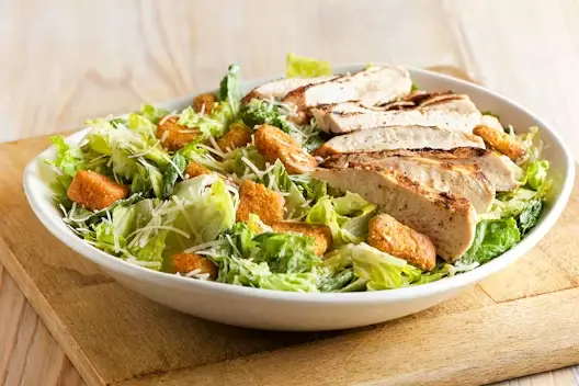 Brisbane Caesar Salad topped with sliced grilled chicken, crunchy croutons, shredded Parmesan, and crisp romaine lettuce served in a white bowl.