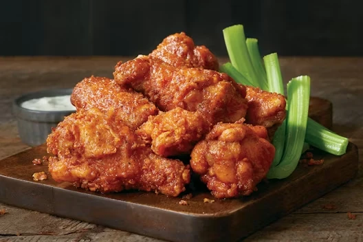 Plate of crispy buffalo chicken wings on a wooden board, served with celery sticks and a cup of dipping sauce in the background.