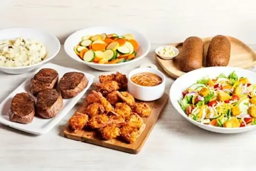 Center-Cut Sirloin & Coconut Shrimp for 4 featuring seasoned sirloin steaks and crispy coconut shrimp served with mashed potatoes, mixed vegetables, fresh garden salad, and dinner rolls with butter on a light wooden table.