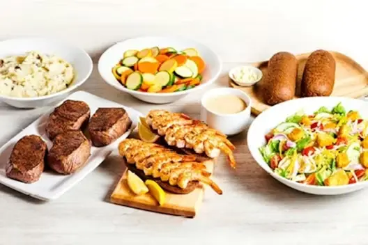 Center-Cut Sirloin & Grilled Shrimp for 4 featuring seasoned sirloin steaks and grilled shrimp skewers served with mashed potatoes, mixed vegetables, fresh garden salad, dinner rolls with butter, and a lemon garnish on a light wooden table.