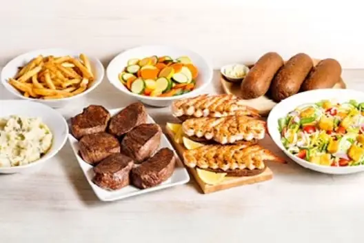 Center-Cut Sirloin & Grilled Shrimp for 6 featuring seasoned sirloin steaks and grilled shrimp skewers, served with mashed potatoes, French fries, mixed vegetables, fresh garden salad, and dinner rolls with butter on a light wooden table.