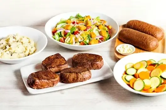 Center-Cut Sirloin for 4 featuring four seasoned sirloin steaks served with mashed potatoes, mixed vegetables, fresh garden salad, and dinner rolls with butter on a light wooden table.