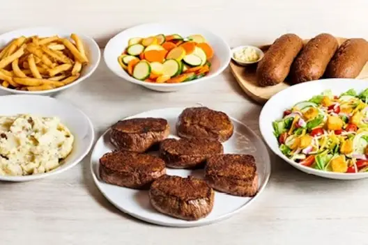 Center-Cut Sirloin for 6 featuring six seasoned sirloin steaks served with mashed potatoes, French fries, mixed vegetables, fresh garden salad, and dinner rolls with butter on a light wooden table.