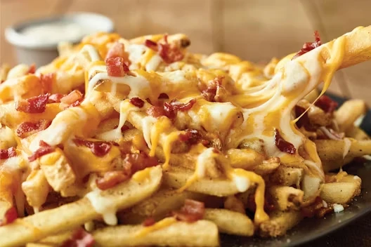 Close-up of a plate of French fries loaded with melted cheese and crispy bacon pieces, with one fry being lifted to show stretchy cheese.