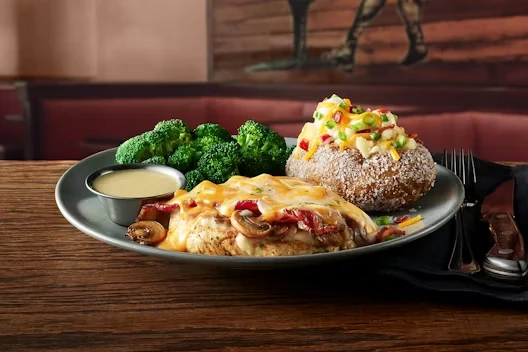 Dinner plate with grilled chicken breast topped with melted cheese, bacon, and mushrooms, served alongside steamed broccoli, a loaded baked potato with cheese and green onions, and a small cup of creamy sauce.