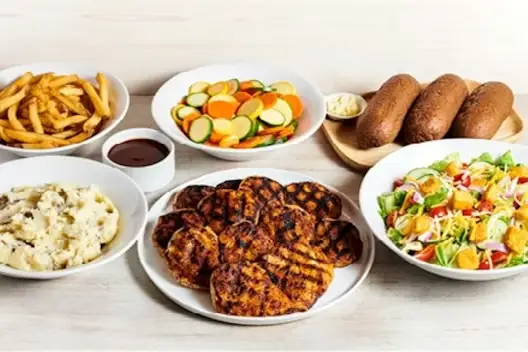 Family-style Grilled Chicken for 6 featuring seasoned grilled chicken breasts, French fries, mashed potatoes, mixed vegetables, fresh garden salad, and bread with butter arranged on a white tabletop.