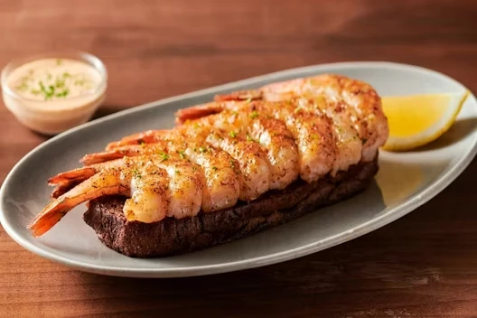 Row of seasoned grilled shrimp arranged on a piece of toasted bread on an oval plate, served with a lemon wedge and a small cup of dipping sauce in the background.