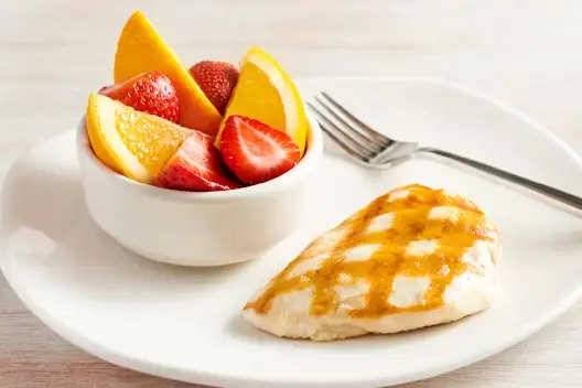 Grilled chicken breast with signature seasoning served with a bowl of fresh fruit including strawberries and orange slices on a white plate.
