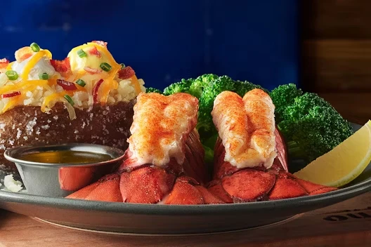 Plate with two lobster tails served alongside a loaded baked potato topped with cheese, bacon, and green onions, steamed broccoli, melted butter in a small cup, and a lemon wedge.