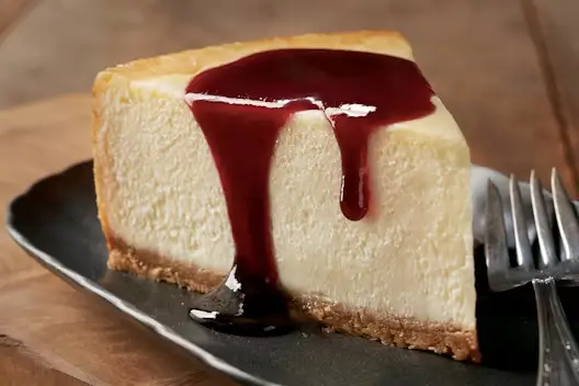 New York-style cheesecake slice topped with rich berry sauce, served on a dark plate with a fork.