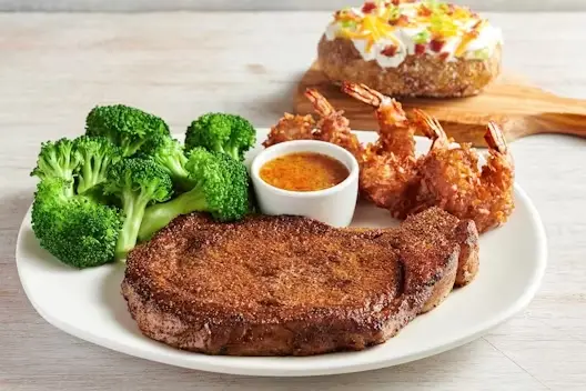 13 oz ribeye steak served with crispy coconut shrimp, steamed broccoli, and a side of dipping sauce on a white dinner plate.