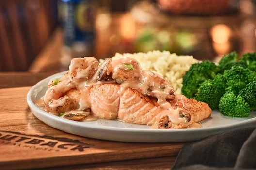 Plate of grilled salmon topped with a creamy mushroom and shrimp sauce, served with steamed broccoli and a side of seasoned rice.