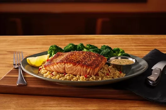 Grilled salmon filet served over seasoned rice pilaf with steamed broccoli, a lemon wedge, and a small cup of creamy herb sauce on the side.