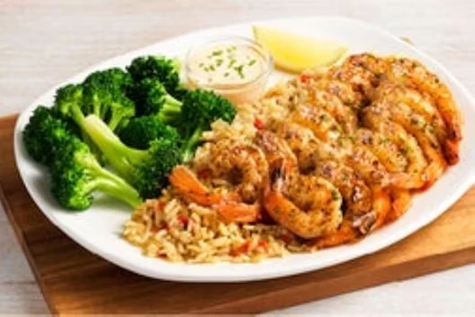 Plate of seasoned grilled shrimp served over rice pilaf with steamed broccoli, a lemon wedge, and a small cup of creamy dipping sauce.