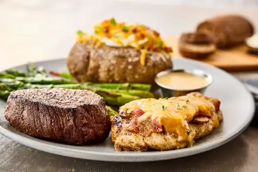 Sirloin steak served with Alice Springs Chicken topped with cheese and bacon, alongside steamed asparagus and a loaded baked potato on a dinner plate.