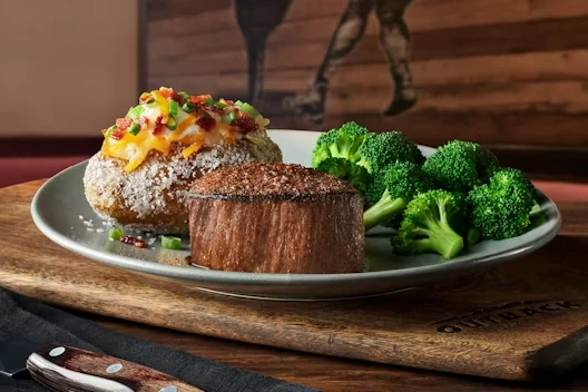 Plate with a seasoned filet steak, a loaded baked potato topped with cheese and bacon, and a side of steamed broccoli, served on a wooden board.